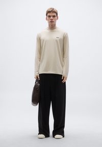 Bézs színű hosszú ujjú ing finom logóval, fekete, túlméretezett nadrággal és krémszínű bebújós cipővel, kezében sötétbarna táskával.