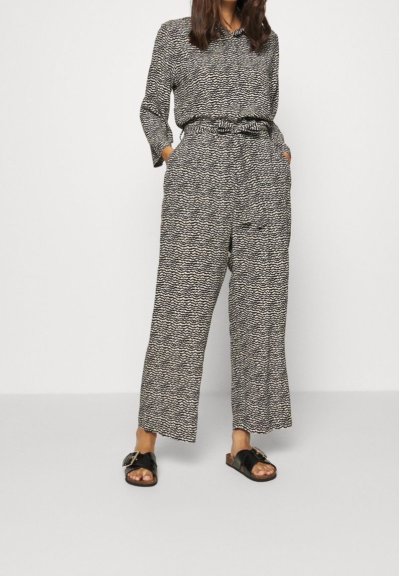 Combinaison à motifs noir et blanc avec manches longues, taille à cordon, jambes larges et poches latérales, associée à des sandales noires à enfiler.