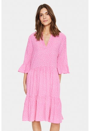 Robe rose avec des pois blancs, décolleté en V, manches trois-quarts évasées et jupe à volants. Confectionnée en tissu léger.