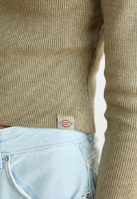 Żebrowany sweter w oliwkowym kolorze z boku, w połączeniu z jasnoniebieskimi dżinsami, prezentujący swobodny i nowoczesny design.