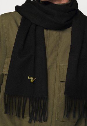 Écharpe noire à franges avec logo Barbour jaune portée sur une veste vert olive avec poches à rabat sur la poitrine.