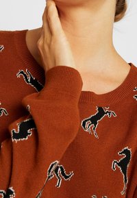 Pull en tricot marron avec un motif de chevaux noirs. Texture douce, poignets côtelés et encolure ronde classique.