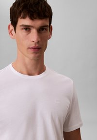 Biała bawełniana koszulka z okrągłym dekoltem i krótkimi rękawami, z subtelnym haftowanym logo na lewej piersi. Gładka tekstura, minimalistyczny design.