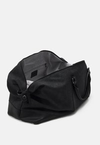 Sac de sport noir avec un extérieur en tissu texturé et une base en cuir lisse. Le compartiment principal zippé révèle une doublure rayée et une poche intérieure.