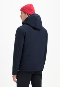 Marineblauwe jas met capuchon, met een gladde textuur en subtiele stikdetails. Gecombineerd met een heldere rode gebreide muts.