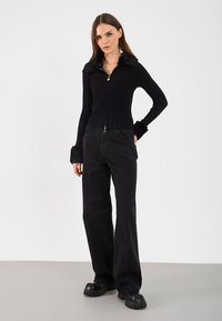 Czarna prążkowana bluza na zamek z kołnierzem i mankietami z futerka, zestawiona z czarnymi spodniami o szerokich nogawkach oraz masywnymi czarnymi butami na platformie.