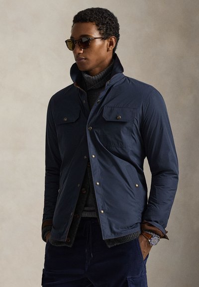 Σκούρο μπλε μπουφάν ελαφρού βάρους με δύο τσέπες στο στήθος, κλείσιμο με κόπιτσες και φερμουάρ στον γιακά. Φορώντας πάνω από γκρι πλεκτό πουλόβερ.