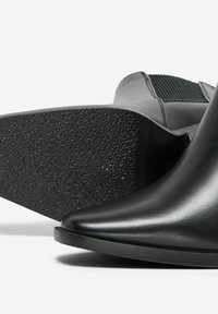 Bottine noire à bout pointu avec une tige en cuir lisse et un talon en caoutchouc texturé. Le talon présente une base plate et solide pour plus de stabilité.