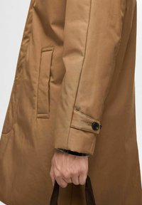 Cappotto marrone con una texture liscia, dotato di una tasca laterale e di un polsino con bottoni. Una persona sta tenendo una borsa marrone.