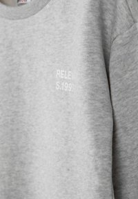 Grå sweatshirt i mjukt, strukturerat tyg. Har rund hals och vittryckt text "RELEVANT S.1991" på framsidan.