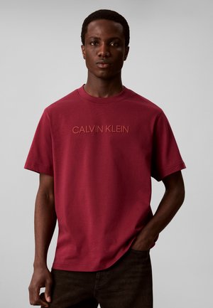 Άνδρας που φοράει κόκκινο T-shirt Calvin Klein με κοντά μανίκια, στέκεται μπροστά σε μονόχρωμο ανοιχτογκρι φόντο.