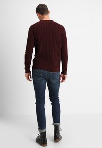 Burgunderfarget strikkegenser med ribbestruktur, iført mørkeblå jeans og svarte ankelstøvler, sett fra bakgrunn mot en hvit bakgrunn.