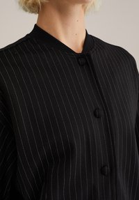 Črna jopica z robom z okroglim ovratnikom. Ima dva okrogla gumba in gladko teksturo z belimi šivi, ki poudarjajo kontrast.