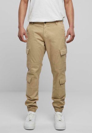 Pantalons cargo beige en coton, avec des poches de chaque côté, une coupe décontractée et des poignets élastiques aux chevilles. Associés à des baskets blanches.