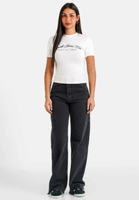 Hvid kortærmet t-shirt med sort print, parret med højtaljede sorte vide jeans og mørke sneakers. Glat stof, afslappet pasform.