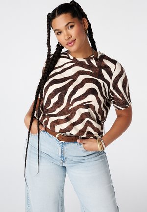 Vrouw met lange vlechten draagt een bruin-wit zebraprintshirt, lichtblauwe jeans, bruine riem en gouden armbanden, hand in haar zak.