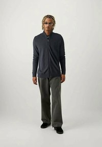 Σκούρο γκρι ζακέτα με φερμουάρ και μακριά μανίκια, συνδυασμένη με ανοιχτόχρωμα γκρι παντελόνια και μαύρα υποδήματα. Το μοντέλο έχει κοντά, κυματιστά μαλλιά.