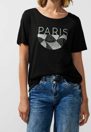 Persoon die een zwart T-shirt draagt met zilverkleurig geometrisch ontwerp en de tekst "PARIS", gecombineerd met blauwe spijkerbroek.