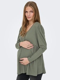 Zwangere vrouw met lang blond haar, gekleed in een losse groene blouse, zachtjes haar buikje vasthoudend terwijl ze naar de camera kijkt.