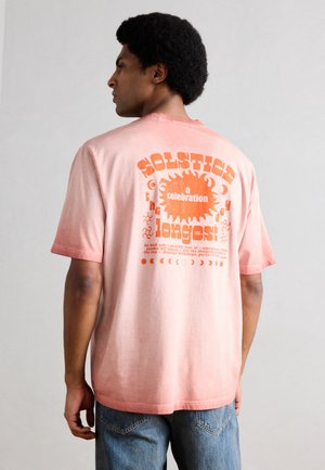 Mann in pinkem Farbverlauf-T-Shirt mit orangefarbenem Sonnen- und Mondmotiv und Text über die Sonnenwendenfeier, trägt blaue Jeans, seitlich stehend.