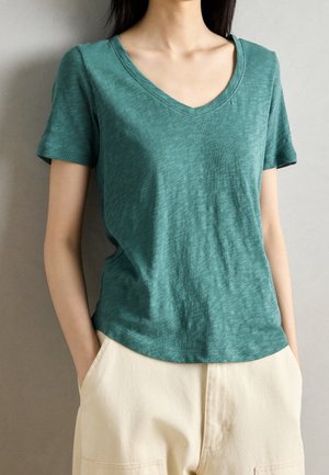 Kobieta ubrana w zielony, krótkim rękawem, T-shirt z dekoltem w serek, z rękami w kieszeniach beżowych spodni z wysokim stanem na jednolitym tle.