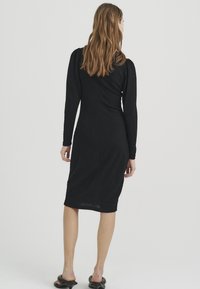 Schwarzes langärmliges Kleid mit figurbetonter Silhouette, strukturiertem Stoff und knielangem Saum. Glatter Rücken ohne auffällige Akzente oder Muster.