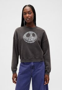 Tmavě šedá krátká mikina s úsměvem vytvořeným z malých stříbrných lebek. Má žebrované manžety a značkový detail na rukávu.