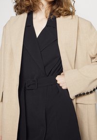 Béžový blazer s texturovanou tkaninou, nošený přes černé šaty s páskem. Výrazné černé knoflíky na rukávech a vrstvený límec.