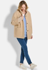 Beige wollen blazer in een gebreid visgraatpatroon, met drie knopen en twee voorkruiken, gecombineerd met blauwe jeans en sneakers.