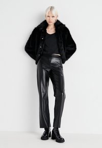 Chaqueta de piel sintética negra con gran cuello, blusa negra ajustada, pantalones de cuero negro brillantes y botines de tacón brillantes.