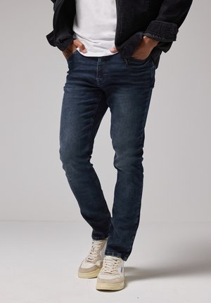 Hombre con jeans azul oscuro, zapatillas blancas, camiseta blanca y chaqueta negra, con las manos en los bolsillos, de pie sobre un suelo blanco.