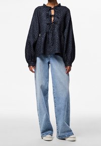 Blouse bleu foncé texturée avec manches bouffantes et ouverture en forme de goutte, associée à un jean large bleu clair et des baskets blanches.