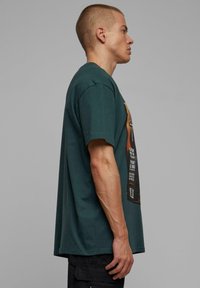 Mørkegrøn bomulds t-shirt med korte ærmer, som har en grafisk print på forsiden, afslappet pasform og rund halsudskæring, der viser fed tekst og billeder.