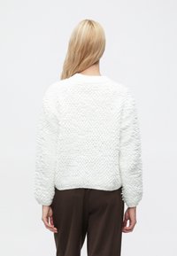 Valkoinen fleeceneule, jossa on teksturoitu, kuoppainen pinta ja pitkät hihat. Pyöreä pääntie ja joustava vyötärönauha. Yhdistetty ruskeisiin housuihin.