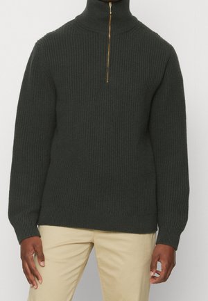 Pull en maille côtelée gris foncé avec col à tirette quart de fermeture dorée, porté avec un pantalon beige par une personne debout.