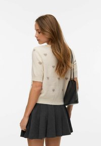 Crèmekleurige trui met korte mouwen en textuurrijke hartpatronen, gecombineerd met een donkergrijze geplooide rok. Gemaakt van een lichtgewicht, zacht materiaal.