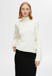 Balta turtlenecka džemperis ar garām piedurknēm un ribotu tekstūru, kombinēts ar melnu svārku. Džemperim ir brīvs griezums un tas ir līdz gurniem.