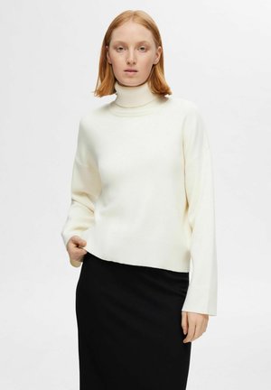 Maglione bianco a collo alto con maniche lunghe e texture a coste, abbinato a una gonna nera. Il maglione ha una vestibilità rilassata e raggiunge l'altezza dell'anca.