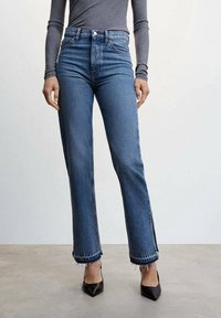 High-waisted blue jeans in a straight-leg cut, featuring frayed hems and side slits, made from denim fabric. Paired with black pointed heels.