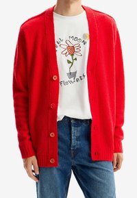 Cardigan rosso lavorato a maglia con vestibilità ampia, dotato di scollatura a V, chiusura con bottoni e polsini a coste. Indossato sopra una t-shirt bianca con design floreale.
