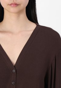 Brun strikket plagg med dyp V-hals, med fire knapper nedover fronten og avslappede lange ermer. Jevn tekstur, uformell design.