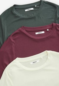 Tre ribbede t-shirts i mørkegrøn, burgunder og off-white. Hver med rund hals og et mærke med teksten "NEXT."