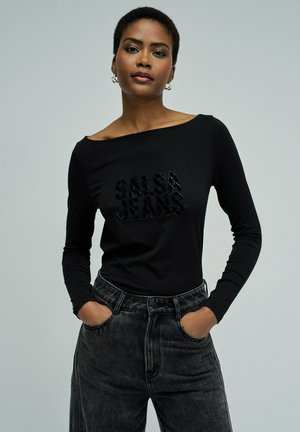 Mujer con camiseta negra de manga larga con texto "SALSA JEANS" y jeans negros descoloridos de tiro alto, manos en los bolsillos, fondo neutro.