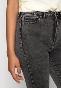 Primer plano de la cintura de una persona, mostrando jeans de mezclilla negros y una camisa de color claro, con una mano descansando junto a la cadera.