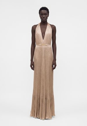 Γυναίκα που φοράει αμάνικο, μακρύ μέχρι το πάτωμα μεταλλικό χρυσό φόρεμα halter με βαθύ V-λαιμόκοψη και λεπτή ζώνη στη μέση, στέκεται μπροστά σε απλό λευκό φόντο.