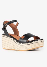 Sandalias de cuña de cuero negro con correa de tobillo, plataforma de yute y plantilla marrón claro con un pequeño parche de logo. Costuras texturizadas en negro como acento.