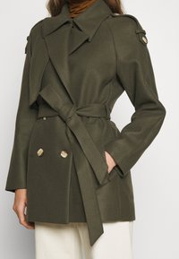 Trench coat verde oliva hecho de tejido texturizado, con un diseño de doble botonadura, amplio cuello, cintura ceñida con cinturón y detalles de botones dorados.