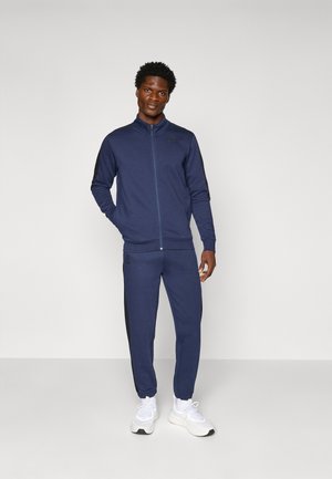 Survêtement en bleu marine en tissu doux ; la veste présente un col haut et une fermeture éclair complète, tandis que le pantalon a des accents noirs sur les côtés et des chevilles resserrées.