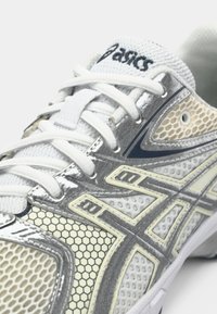 Buty sportowe ASICS w białym i szarym kolorze z siateczkową cholewką, akcentami w kształcie heksagonów, wyściełanym językiem i białymi sznurowadłami.