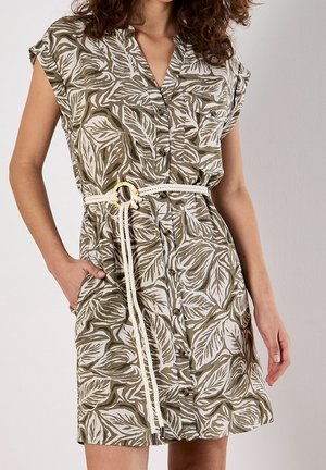 Donna che indossa un vestito con maniche corte e bottoni sul davanti, con motivo di foglie verdi e bianche, fissato da una cintura a corda bianca, una mano in tasca.